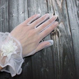 May include: White lace gloves with a ruffled wrist cuff and a white flower embellishment.