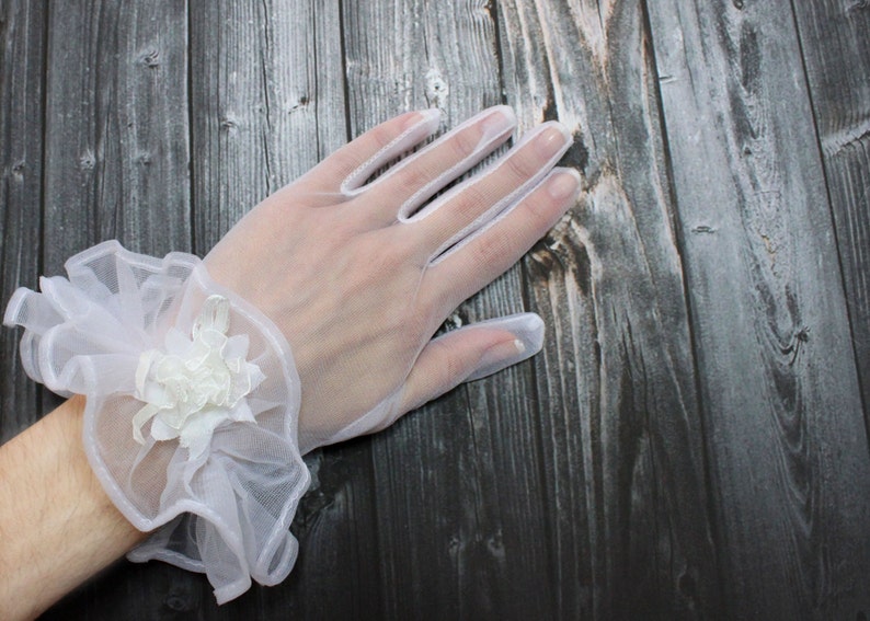 May include: White lace gloves with a ruffled wrist cuff and a white flower embellishment.