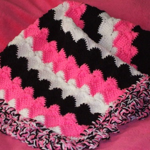 May include: A pink, black, and white crocheted baby blanket with a ruffled edge.