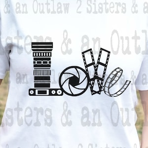 May include: White t-shirt with a black graphic design of a camera, film reel, and lens forming the word "love". The text "2 Sisters & an Outlaw" is repeated around the graphic.