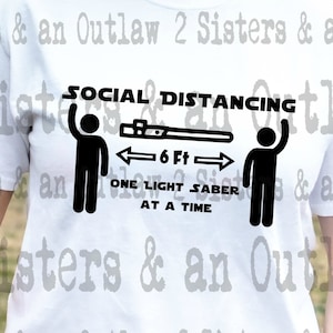 May include: White t-shirt with black text that reads "Social Distancing" and shows two stick figures with a lightsaber between them. The text below the figures reads "One Light Saber At A Time".