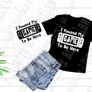 May include: Two t-shirts with the text "I Paused My GAME To Be Here" and a graphic of a video game controller. One shirt is white with black text and the other is black with white text.