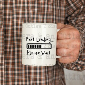Op de afbeelding: Witte keramische mok met de tekst "Fart Loading... Please Wait" in het zwart. De mok heeft een laadbalkgrafiek. De mok wordt vastgehouden door een persoon die een bruin en oranje geruit overhemd draagt.