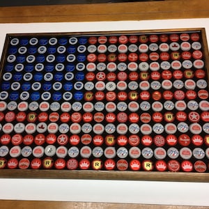 May include: A framed American flag made from bottle caps. The flag is red, white, and blue. The bottle caps are arranged in a grid pattern. The frame is made of wood.
