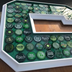 Puede incluir: Una gran letra 'G' enmarcada en blanco, llena de tapas de botellas de cerveza. Las tapas son verdes, plateadas y negras, con nombres de marcas visibles. El fondo es una superficie verde y el marco es blanco.