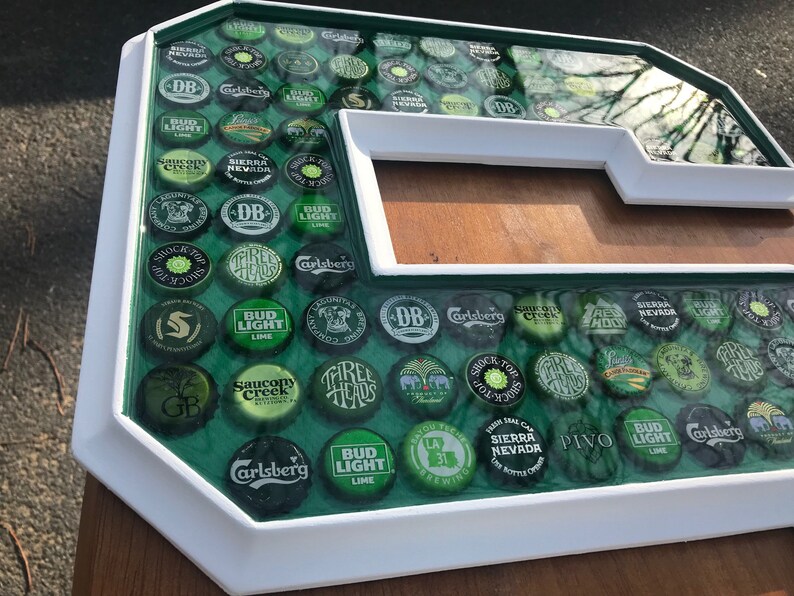 Puede incluir: Una gran letra 'G' enmarcada en blanco, llena de tapas de botellas de cerveza. Las tapas son verdes, plateadas y negras, con nombres de marcas visibles. El fondo es una superficie verde y el marco es blanco.