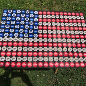 May include: A black surface covered with bottle caps arranged in the pattern of the American flag. The top row of the flag is blue and features Bud Light bottle caps. The middle row is white and features Miller Lite bottle caps. The bottom row is red and features Miller Lite bottle caps.