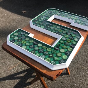 Puede incluir: Una gran letra "S" con marco blanco, llena de tapas de botellas verdes. Las tapas son visibles a trav&eacute;s de una superficie transparente te&ntilde;ida de verde. La letra se muestra sobre una mesa de madera, mostrando un dise&ntilde;o decorativo &uacute;nico.