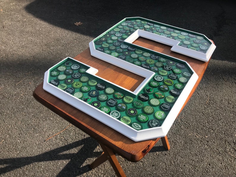 Puede incluir: Una gran letra "S" con marco blanco, llena de tapas de botellas verdes. Las tapas son visibles a trav&eacute;s de una superficie transparente te&ntilde;ida de verde. La letra se muestra sobre una mesa de madera, mostrando un dise&ntilde;o decorativo &uacute;nico.