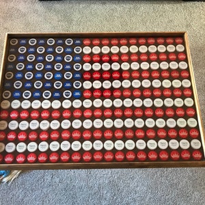 Puede incluir: Una mesa de madera con tapa de mesa con un mosaico de tapas de botellas rojas, blancas y azules dispuestas en forma de bandera estadounidense.