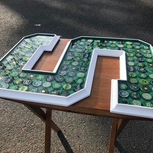 Puede incluir: Grandes letras "M" y "S" con marco blanco, llenas de tapas de botellas verdes. Las letras tienen un borde verde y resina transparente. Las letras est&aacute;n sobre una mesa de madera. El fondo es una superficie pavimentada.