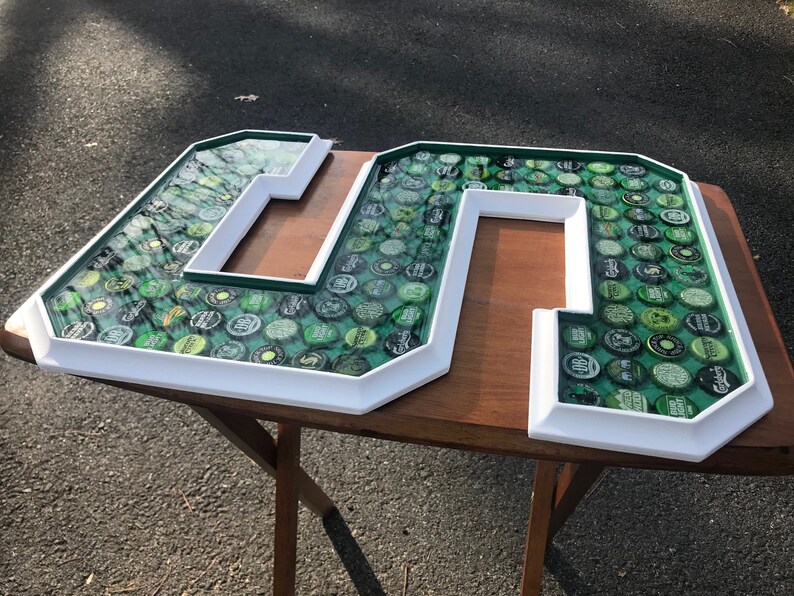 Puede incluir: Grandes letras "M" y "S" con marco blanco, llenas de tapas de botellas verdes. Las letras tienen un borde verde y resina transparente. Las letras est&aacute;n sobre una mesa de madera. El fondo es una superficie pavimentada.