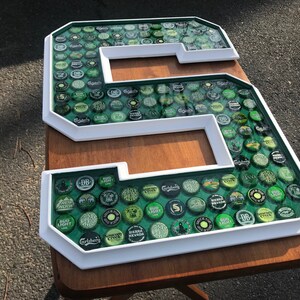 Puede incluir: Una gran letra "S" con marco blanco, llena de varias tapas de botellas verdes y plateadas. Las tapas est&aacute;n colocadas sobre un fondo verde y encapsuladas en una resina transparente. La letra se muestra sobre una superficie de madera.