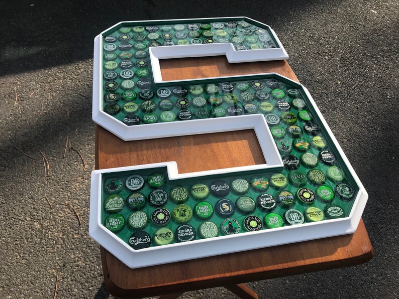 Puede incluir: Una gran letra "S" con marco blanco, llena de varias tapas de botellas verdes y plateadas. Las tapas est&aacute;n colocadas sobre un fondo verde y encapsuladas en una resina transparente. La letra se muestra sobre una superficie de madera.