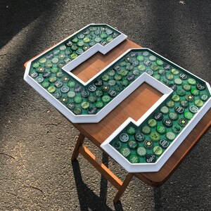 Puede incluir: Un gran n&uacute;mero "5" enmarcado en blanco, lleno de tapas de botellas verdes y plateadas. Las tapas est&aacute;n colocadas sobre un fondo verde. La obra de arte se exhibe sobre una peque&ntilde;a mesa de madera marr&oacute;n, al aire libre.