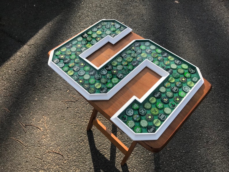 Puede incluir: Un gran n&uacute;mero "5" enmarcado en blanco, lleno de tapas de botellas verdes y plateadas. Las tapas est&aacute;n colocadas sobre un fondo verde. La obra de arte se exhibe sobre una peque&ntilde;a mesa de madera marr&oacute;n, al aire libre.