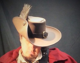 May include: A tan wide-brimmed hat with a dark brown band, a brass buckle, and a single feather accent. The hat is worn with a cream-colored shirt, a red coat with brass buttons, and a dark leather shoulder strap featuring brass studs and a buckle.