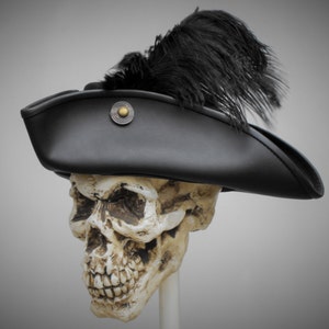 May include: A black tricorn hat featuring a large black feather plume attached to the side and a circular metallic button with a brass-coloured centre detail. The hat is positioned on a realistic human skull model against a neutral background.