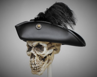 May include: A black tricorn hat featuring a large black feather plume attached to the side and a circular metallic button with a brass-colored center detail. The hat is positioned on a realistic human skull model against a neutral background.