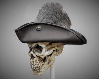Puede incluir: Un sombrero de tres picos de color marrón oscuro con una pluma gris grande y erguida y un botón de metal color latón en el lateral. El sombrero se muestra sobre un modelo de cráneo humano realista, destacando el ala curva y su estructura.