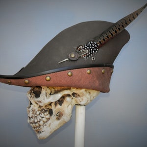 May include: A brown leather tricorn hat with a feather accent and brass studs. The hat is on a skull mannequin.