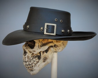Puede incluir: Un sombrero vaquero de cuero negro con ala ancha y hebilla plateada. El sombrero está decorado con pequeños tachuelas de calavera plateadas.