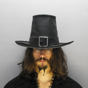 May include: A black tall-crowned hat featuring a wide brim and a black hatband adorned with a prominent silver-toned rectangular buckle. The hat has a structured, cylindrical crown and a flat brim that extends horizontally from the base.