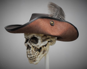 May include: A brown tricorn hat with a textured suede-like finish, featuring a black leather trim along the brim, a single brass-toned metal button accent on the side, and a grey feather plume tucked into the folded brim, resting on a skull.