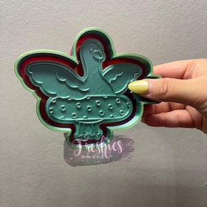 May include: A teal-coloured cookie cutter shaped like a bird with outstretched wings, atop a mushroom. It has a red and green border. The word "Freshies" is printed on the bottom.
