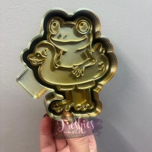 May include: A gold-colored cookie cutter in the shape of a frog wearing a duck floatie. The frog has large eyes and is smiling. The cookie cutter is held by a hand, with the text "Freshies in the U.S.A." visible.