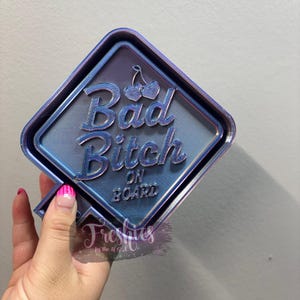 Op de afbeelding: Een blauw en paars bord met de woorden "Bad Bitch ON BOARD" in een gestileerd lettertype. Het bord heeft een ruitvorm met een kersontwerp boven het woord "Bitch". Het bord wordt vastgehouden door een hand met roze en witte nagellak.