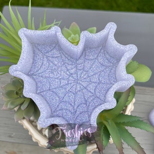 May include: A light purple heart-shaped dish with a spiderweb design. The dish is made of resin and has a glittery finish.