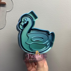 May include: A teal blue and green 3D printed plastic flamingo pool float cookie cutter. The cookie cutter is shaped like a flamingo with a ring around its neck.