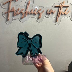 May include: Teal blue silicone mold shaped like a bow. The mold is being held by a hand.