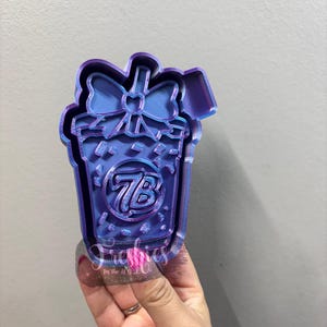 May include: A multi-layered, blue and purple cookie cutter in the shape of a gift box with a bow on top. The gift box has the letters "7B" inside. The text "Freshies" is visible at the bottom.
