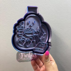 May include: A purple and blue cookie cutter in the shape of a ghost, holding popcorn, a drink, and a book labeled "True Crime." The cutter has a smiling face and a layered design. The text "Freshies" is visible.