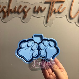 May include: A light blue paw print mold with two paw prints. The mold is held in front of a sign that says "Freshies in the U.S.A."