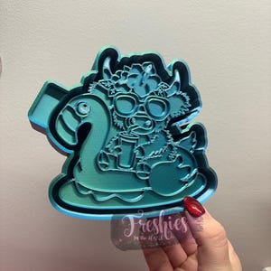May include: A teal blue 3D printed cookie cutter shaped like a cow wearing sunglasses and a flower crown, floating on a blue and white inflatable ring.
