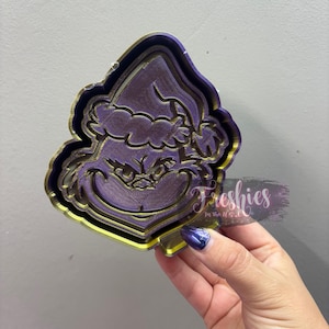 May include: A purple and gold tray with a Grinch design. The tray has a detailed, raised design of the Grinch's face and hat. The edges are outlined in gold. The tray is held by a hand with a purple nail polish.