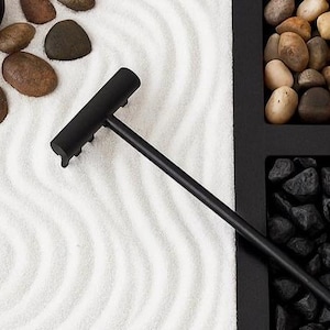 May include: A black zen garden rake with a long handle rests on white sand with wave patterns. The image also shows smooth, brown stones and a black container filled with black rocks and other stones.