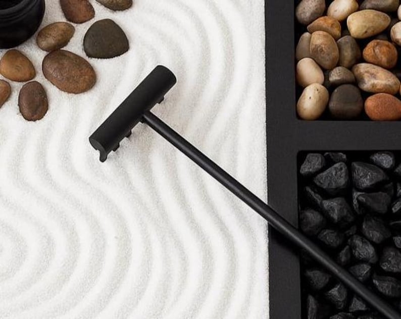 May include: A black zen garden rake with a long handle rests on white sand with wave patterns. The image also shows smooth, brown stones and a black container filled with black rocks and other stones.