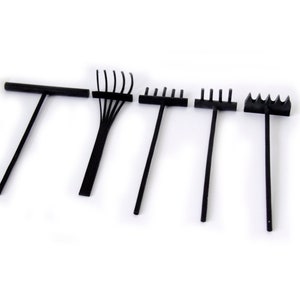 May include: Five black zen garden rakes of varying designs are arranged against a white background. The rakes include a T-shaped rake, a rake with curved tines, and rakes with straight tines. These tools are used for creating patterns in sand gardens.