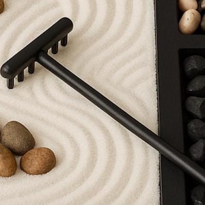May include: A black miniature zen garden rake with a long handle and a row of tines. The rake is positioned on a bed of white sand with curved patterns. Brown and gray stones are scattered around the sand, and black stones are in a black tray.