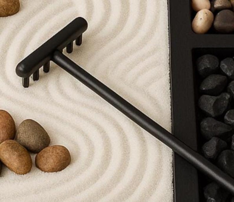 May include: A black miniature zen garden rake with a long handle and a row of tines. The rake is positioned on a bed of white sand with curved patterns. Brown and gray stones are scattered around the sand, and black stones are in a black tray.