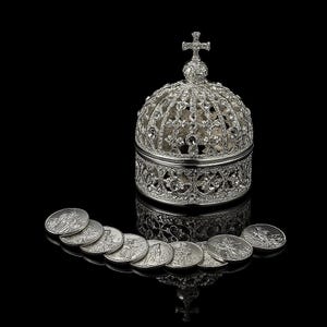 Peut inclure: Une boîte argentée en forme de couronne, surmontée d'une croix, ornée de cristaux décoratifs. Plusieurs pièces de monnaie argentées avec des gravures détaillées sont disposées devant la boîte, sur fond noir.