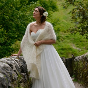 Peut inclure: Une mariée portant une robe de mariée blanche avec un châle en tricot blanc et une fleur blanche dans les cheveux. Elle se tient sur un pont de pierre avec une végétation verte en arrière-plan.