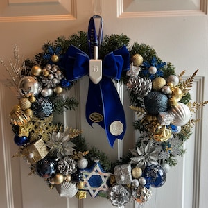 May include: A blue and silver Christmas wreath with a blue velvet bow and a dreidel hanging from the bow. The wreath is decorated with pine cones, snowflakes, and gold and silver baubles.