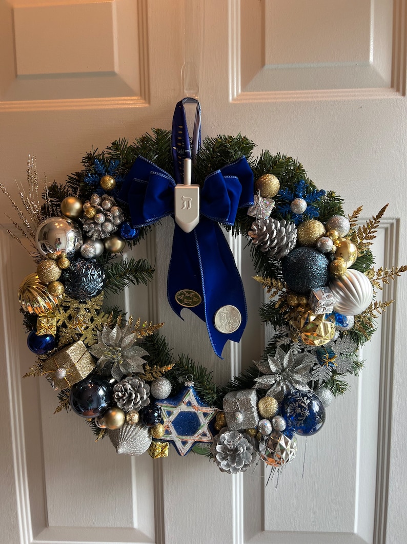 May include: A blue and silver Christmas wreath with a blue velvet bow and a dreidel hanging from the bow. The wreath is decorated with pine cones, snowflakes, and gold and silver baubles.