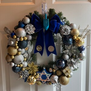 May include: A blue and silver Christmas wreath with a blue velvet bow and a blue dreidel in the centre. The wreath is decorated with gold and silver baubles, pine cones, and sprigs of greenery.