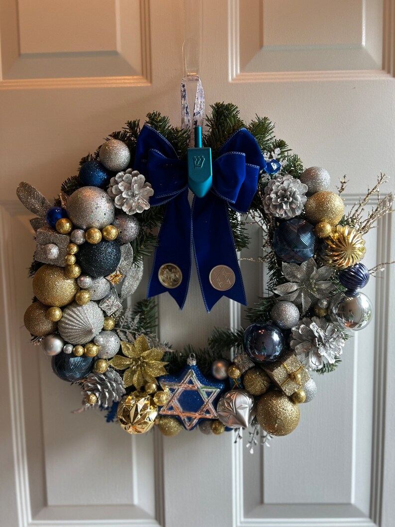 May include: A blue and silver Christmas wreath with a blue velvet bow and a blue dreidel in the centre. The wreath is decorated with gold and silver baubles, pine cones, and sprigs of greenery.